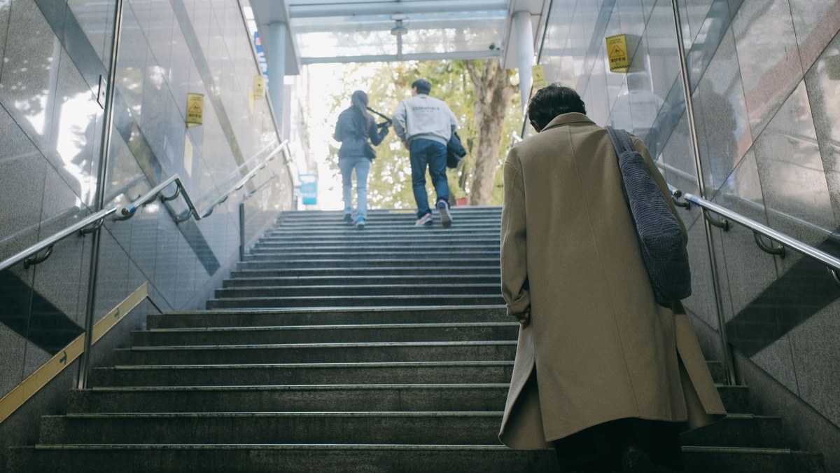 Persona con abrigo y bolso subiendo a pie unas escaleras urbanas con barandal, con luz natural al fondo